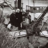 van Rob Bruijn - Het echtpaar Wijkstra op het voordek van hun tjalkje vlak naast de spoorbrug in de Purmerends Where, 1991